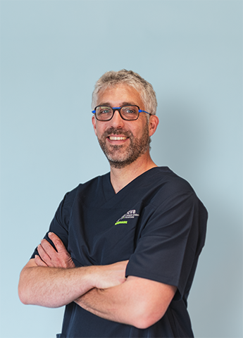 Smiling man with gray hair and glasses wearing navy blue medical scrubs with folded arms against a light blue background.