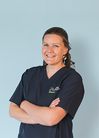 Smiling woman with brown hair in a navy blue medical scrub top standing with arms crossed against a light blue background.
