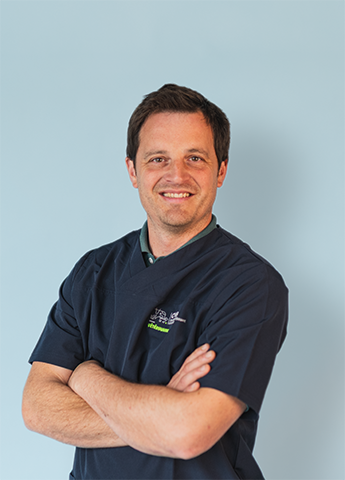 Smiling man in dark medical scrubs with arms crossed against a light blue background.