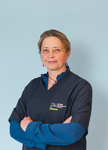 Woman with short hair wearing a navy blue medical uniform and a blue long-sleeve shirt, standing with arms crossed against a light blue background.