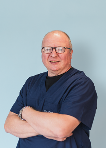 Man in navy blue medical scrubs with glasses and arms crossed standing against a light blue background.
