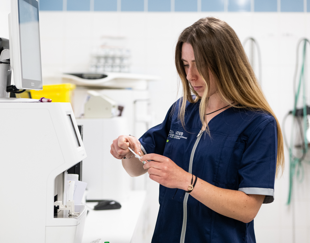Female laboratory technician in navy scrubs preparing a sample near a white analyzer machine in a clinical lab.