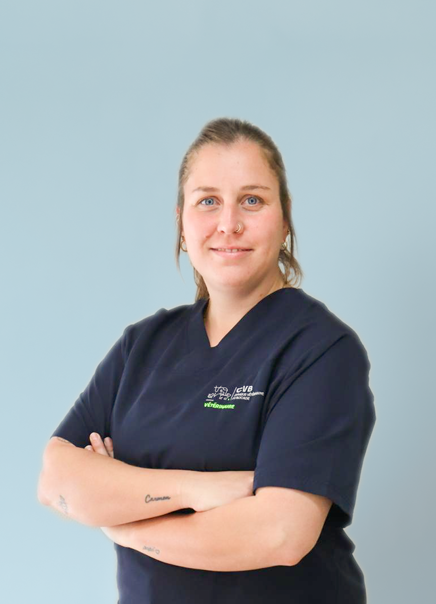 Smiling woman with blue eyes and nose ring wearing dark blue veterinarian scrubs with arms crossed against a light blue background.