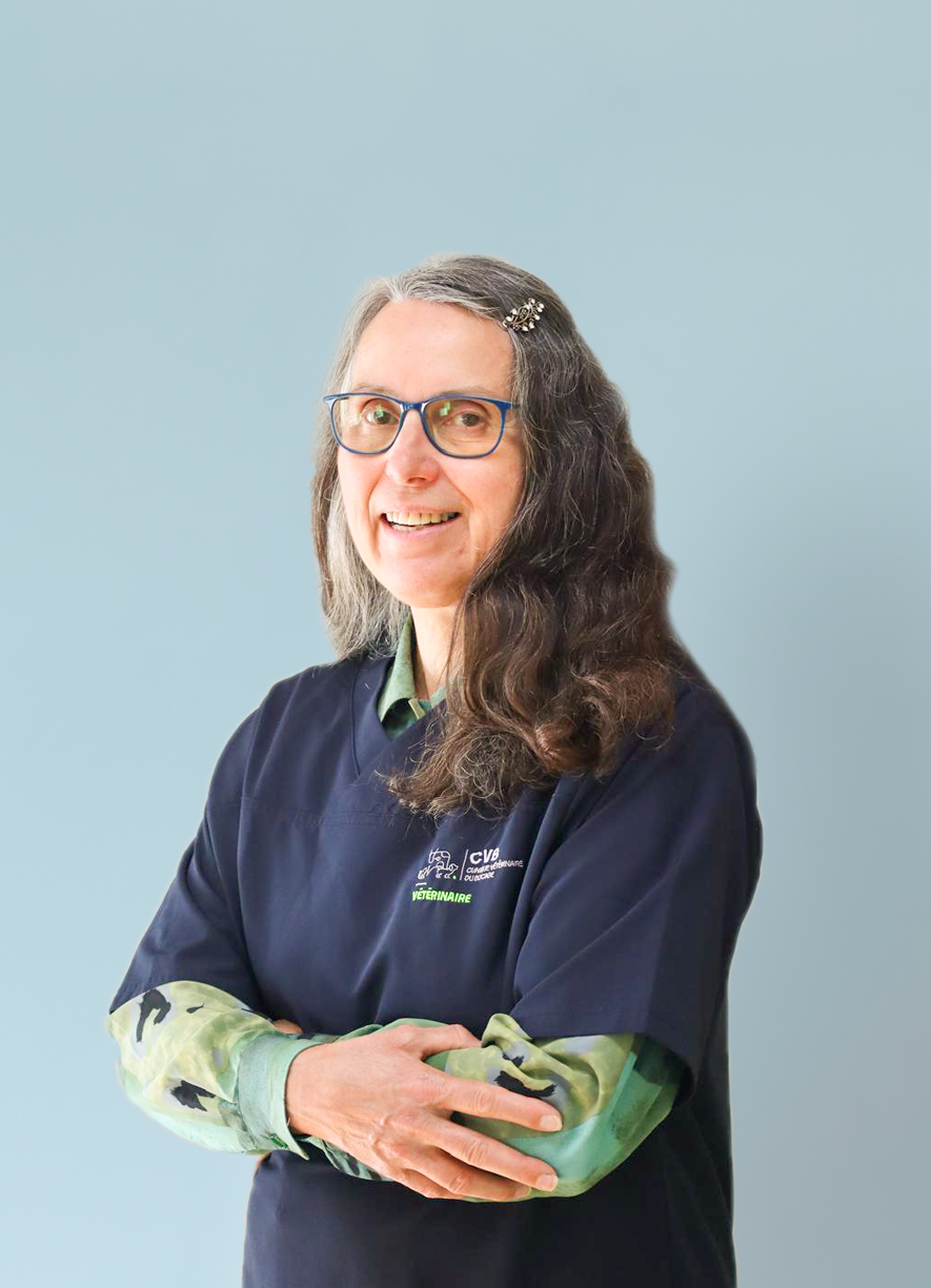Woman with long gray and brown hair, wearing glasses and a dark veterinary uniform, smiling with arms crossed against a light blue background.