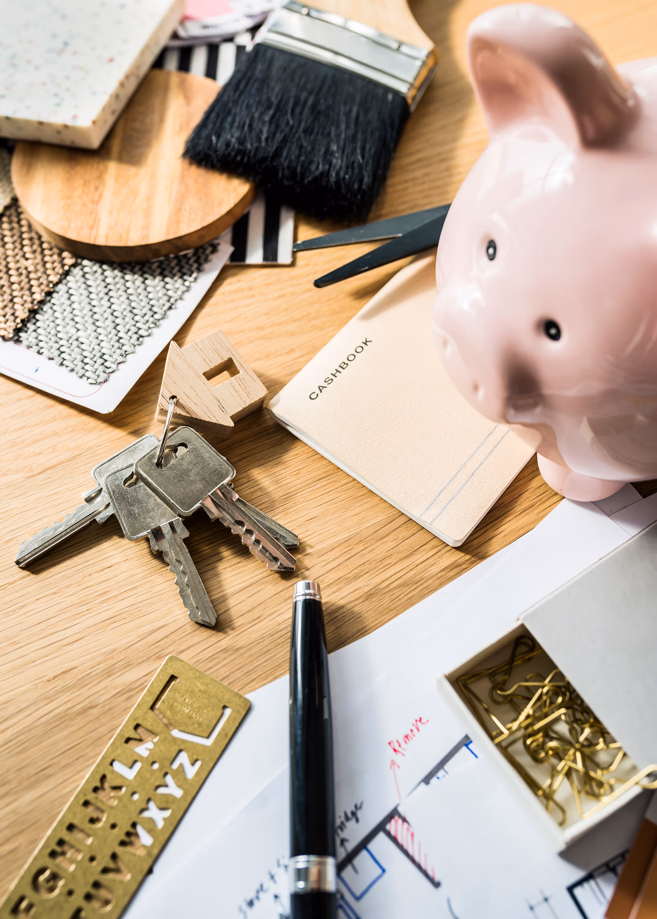 A flat lay of house keys, piggy bank, check books, pens, and paper