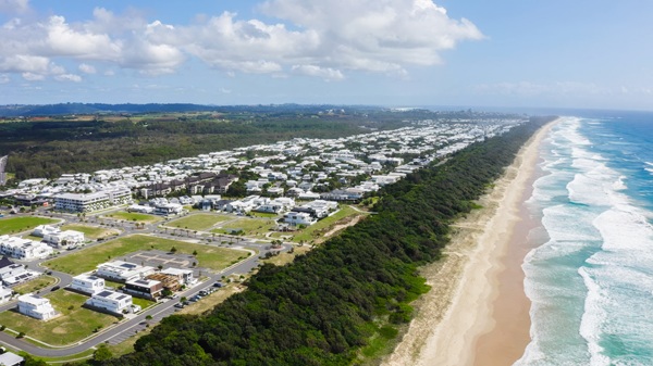 Aerial view of a coastal residential area with white houses, a dense green forest strip, and a long sandy beach with waves under a partly cloudy sky.