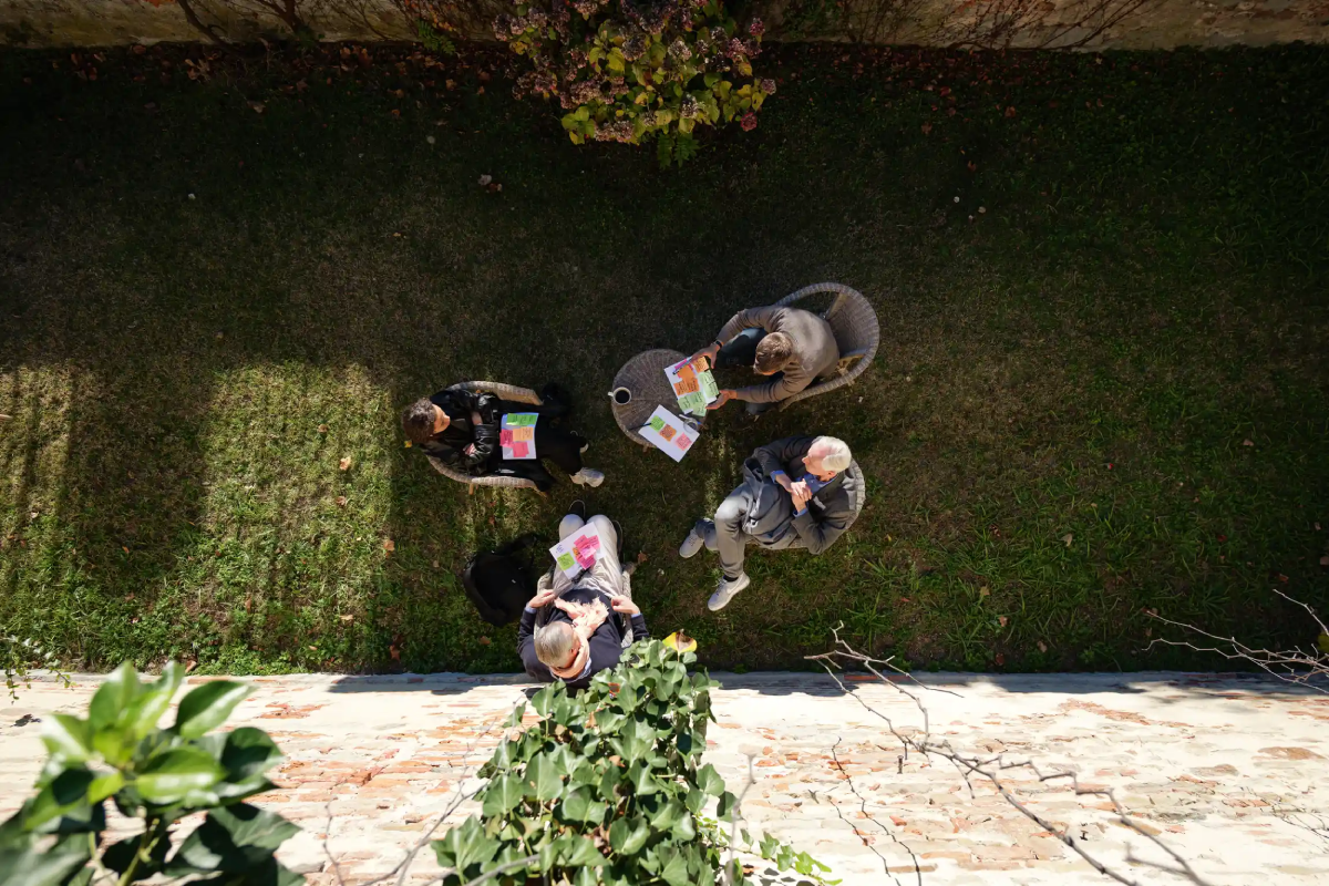 Four people are sitting in a grassy area, discussing while holding colorful papers. They are surrounded by a stone wall and greenery, enjoying a bright, sunny day.
