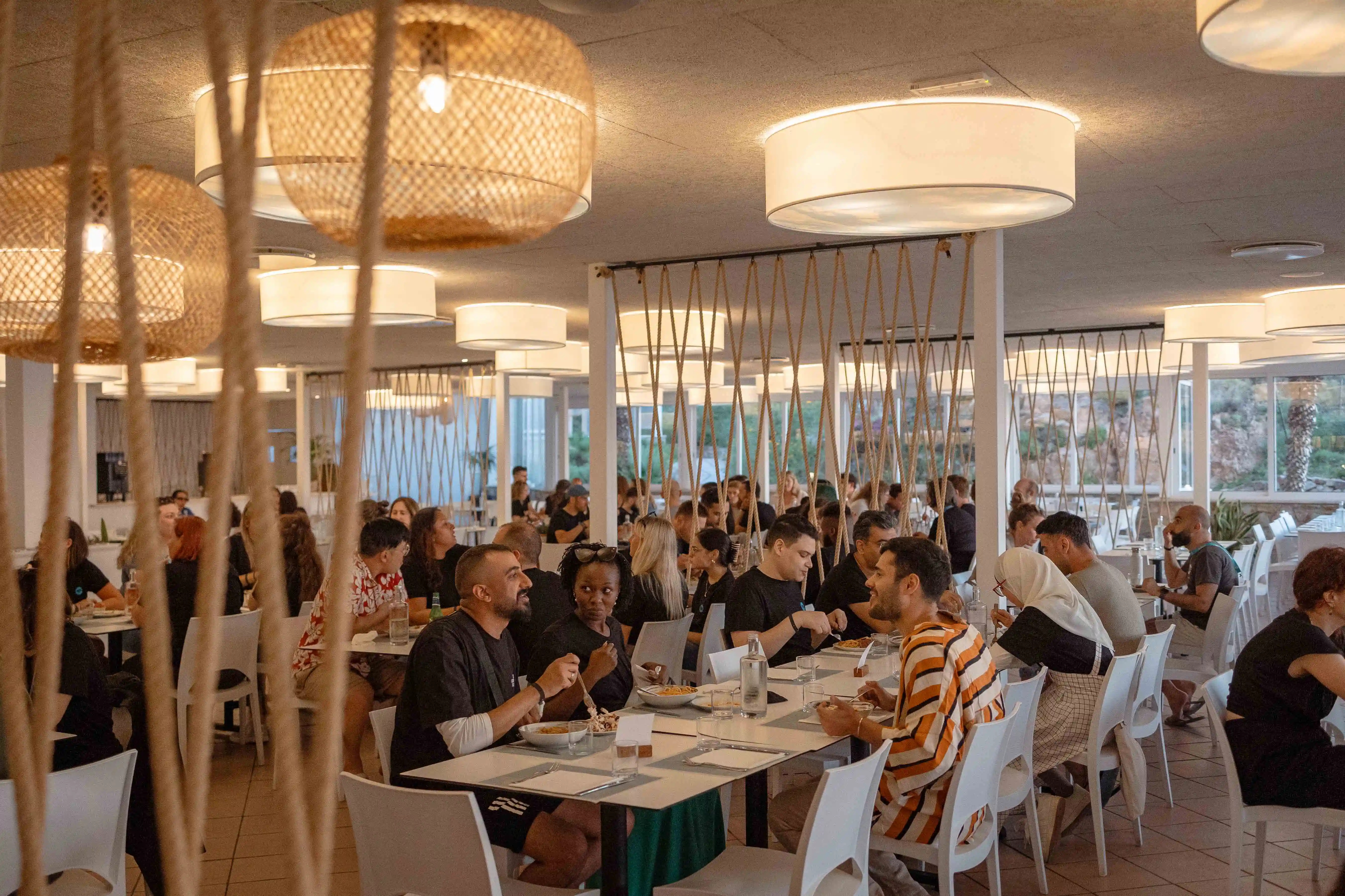 Modern restaurant with many people dining at tables under warm hanging lights and large windows.