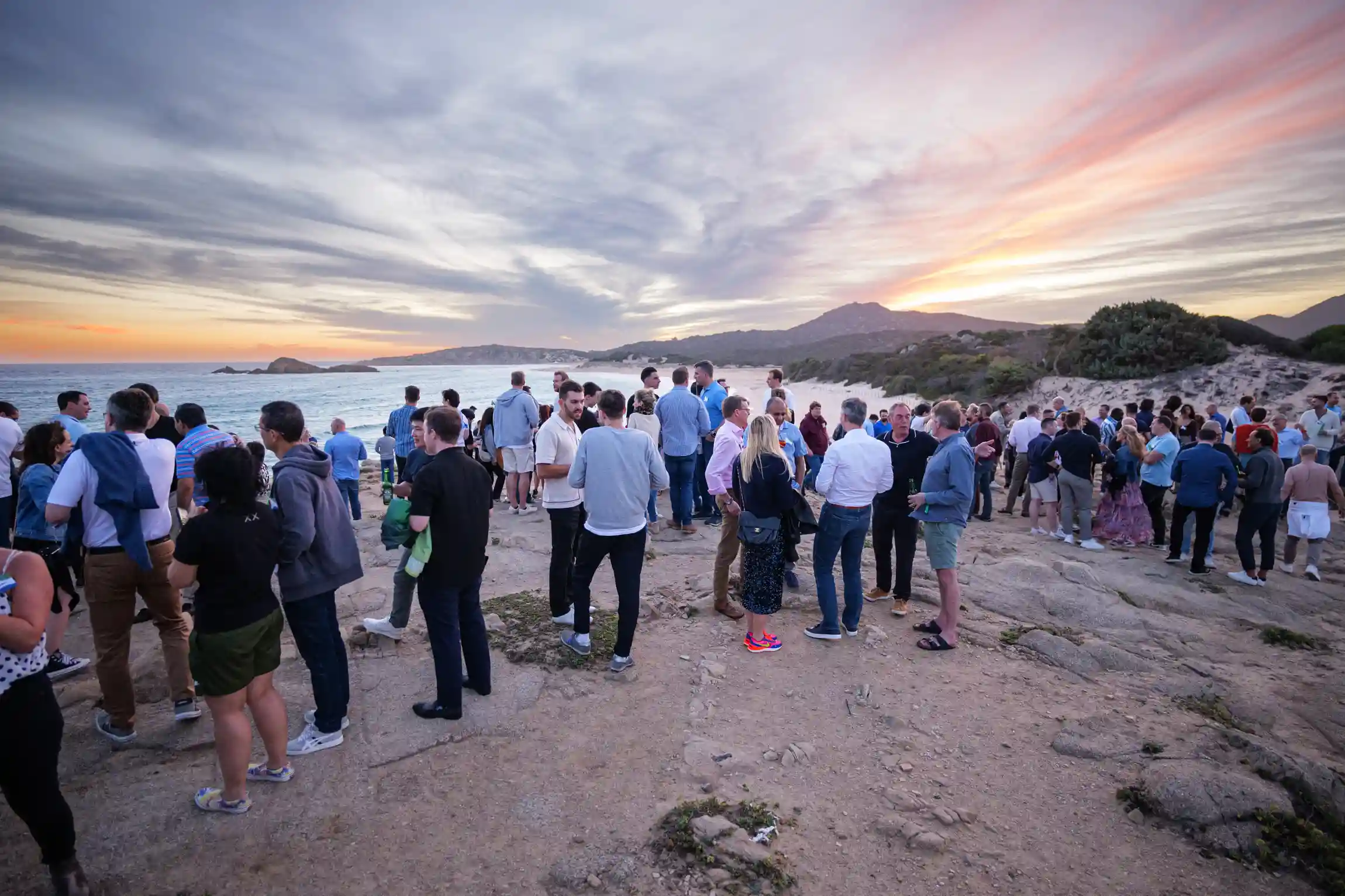 Two individuals sit on grass in a scenic outdoor setting, examining a bottle. The environment features rolling hills and distant trees, with other people relaxing nearby in casual attire.