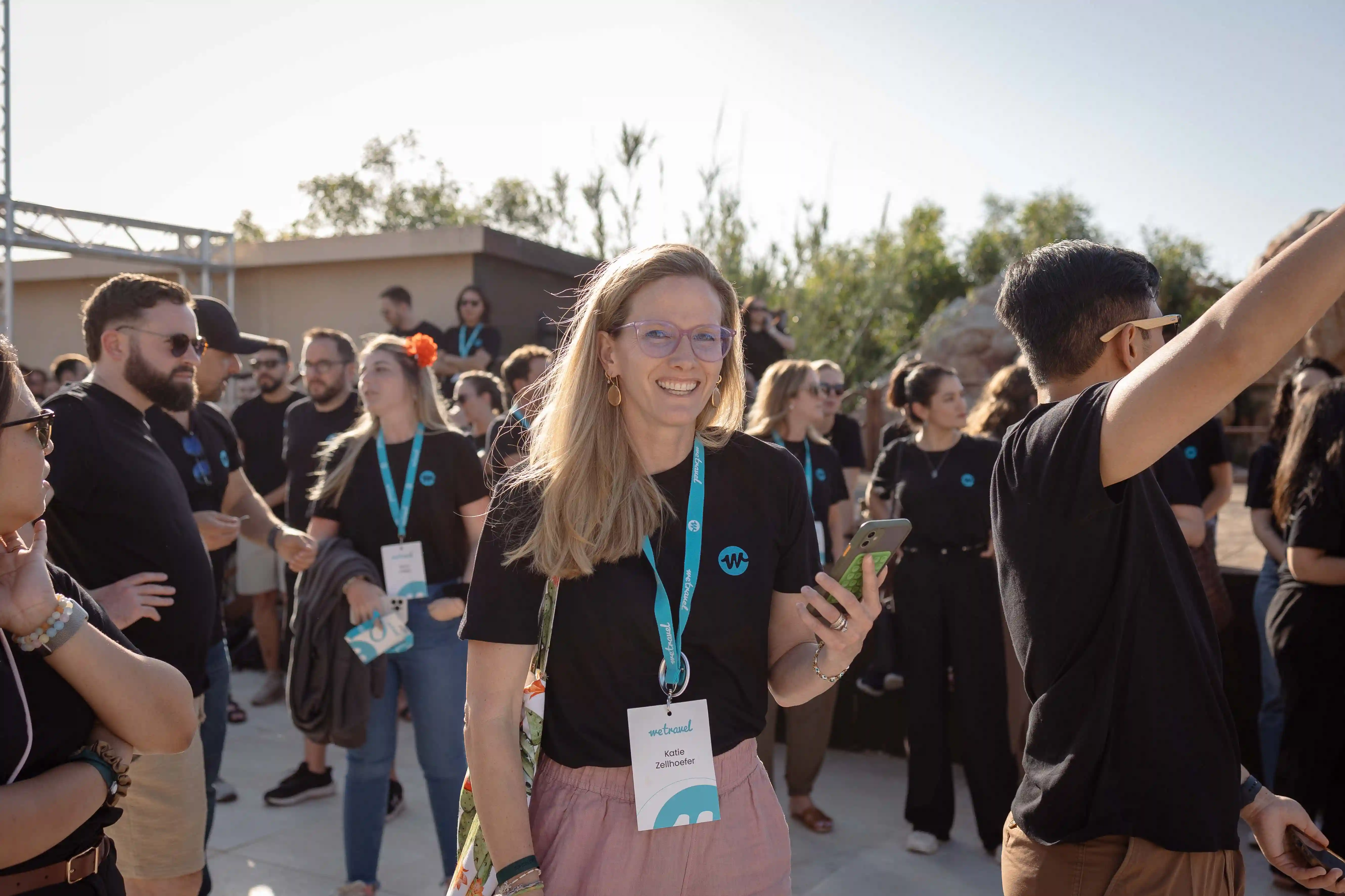 Smiling woman wearing glasses and a conference badge holding a phone among a group of casually dressed people outdoors.