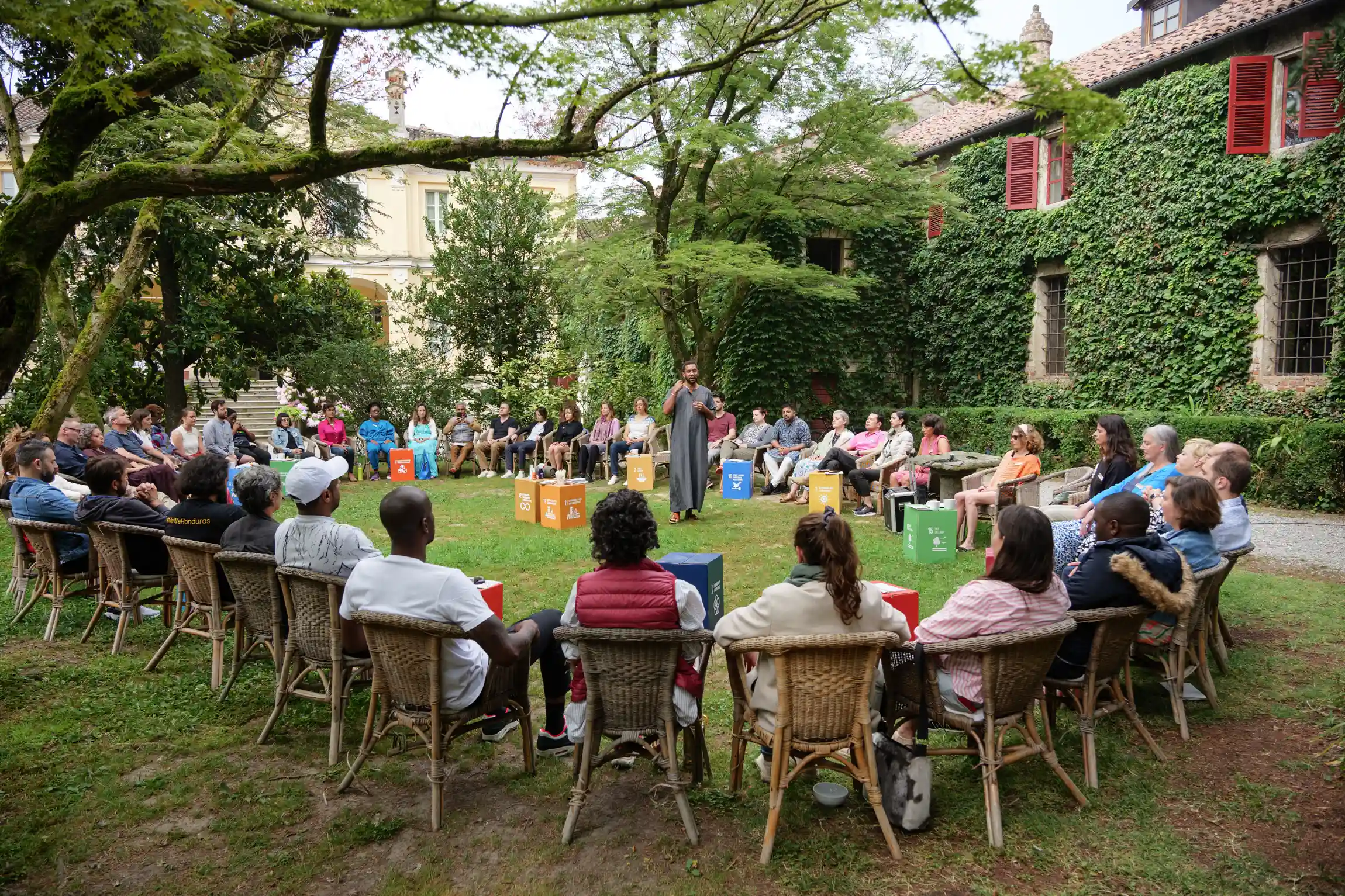 Diverse group of people seated in a large circle outdoors on grass, with a person standing and speaking in the center near color-coded cubes with sustainability icons.
