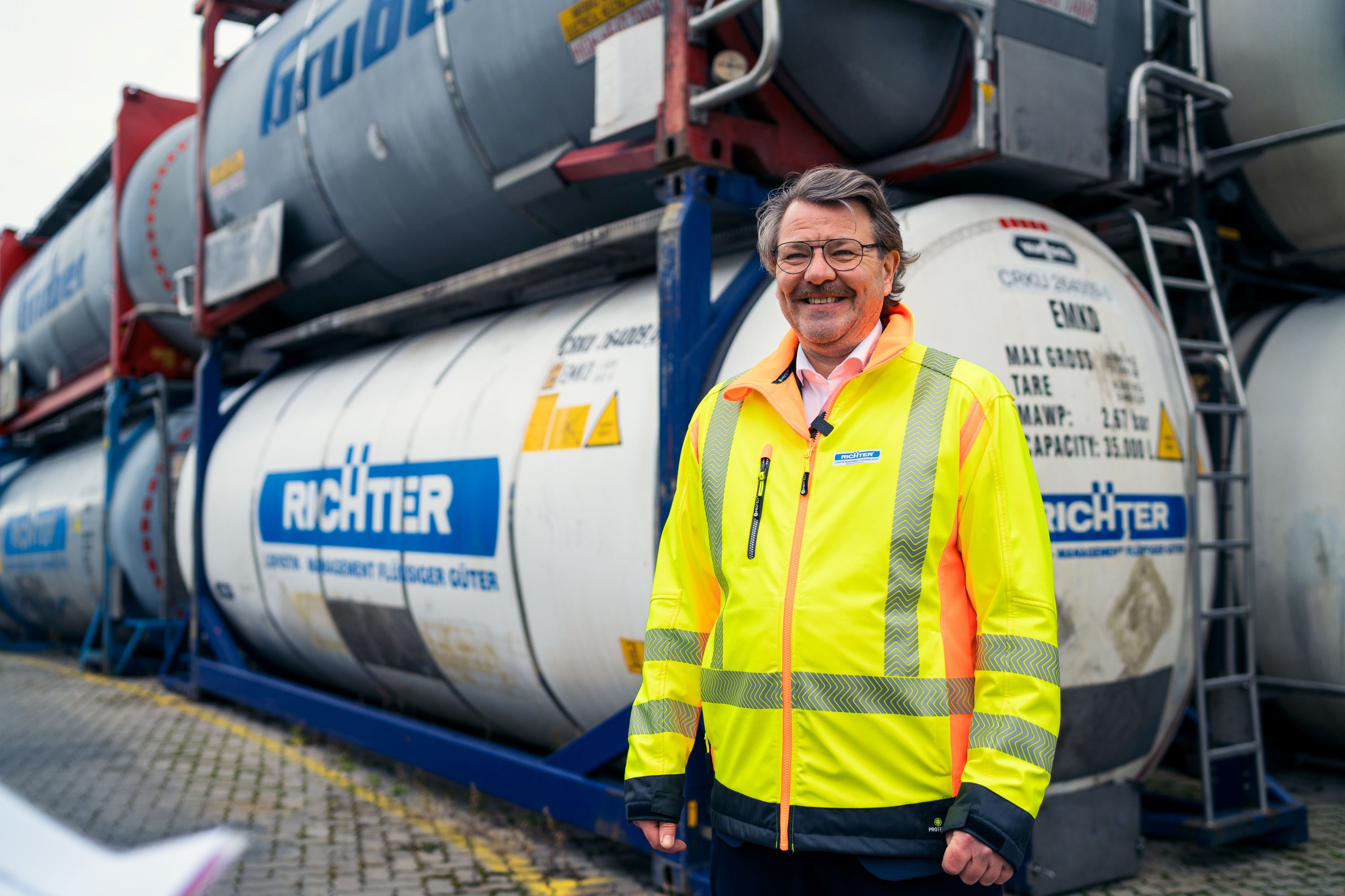 Kurt Richter vor Tankcontainern auf einem Logistikstandort. Das Bild steht für Chemielogistik, Tankcontainermanagement, Supply Chain Collaboration, sichere Gefahrgutprozesse und die effiziente Steuerung komplexer Transportketten. | Kurt Richter in front of tank containers at a logistics site. The image represents chemical logistics, tank container management, supply chain collaboration, safe hazardous goods processes, and the efficient management of complex transportation networks.