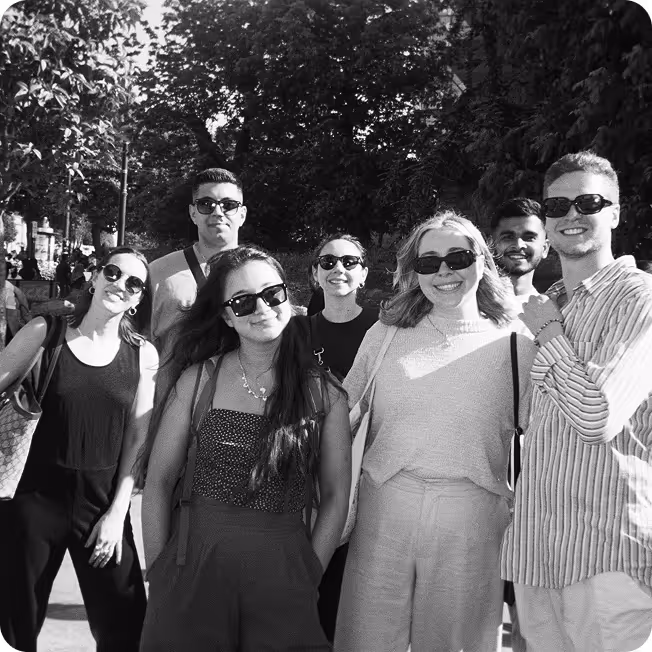 Group of seven young adults smiling and posing outdoors wearing sunglasses in a sunny park setting.