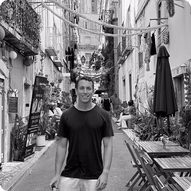 Smiling man in a black t-shirt standing on a narrow street lined with tables, chairs, and plants in a European-style neighborhood.