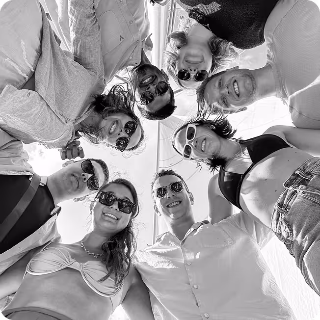 Group of eight young adults wearing sunglasses, smiling and standing closely in a circle looking down at the camera.