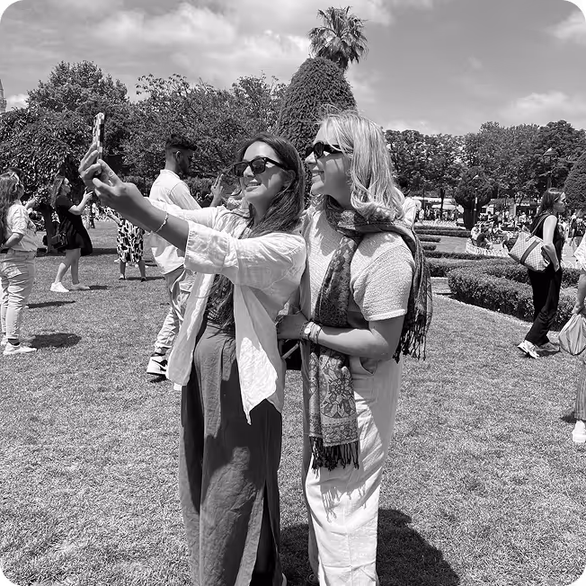 Two women wearing sunglasses smiling and taking a selfie together outdoors in a park with several other people in the background.