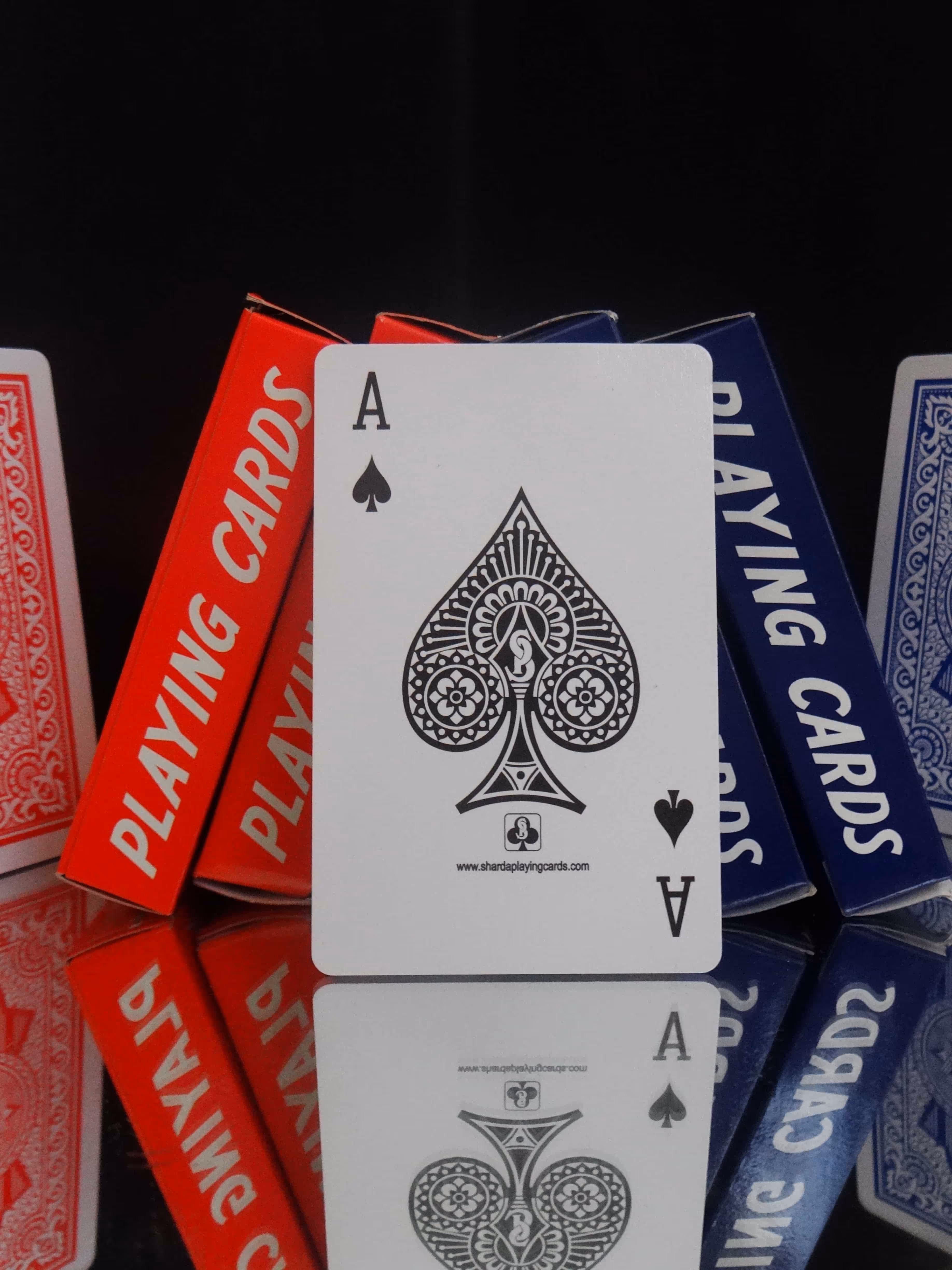 Red and blue Charminar tuck boxes placed side by side with the Ace of Spades in the center, positioned on a mirror surface reflecting the deck’s elegant design and intricate detailing.