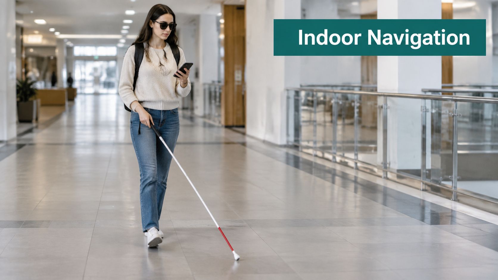 A young woman wearing sunglasses walks through a hallway while using a white mobility cane and smartphone.