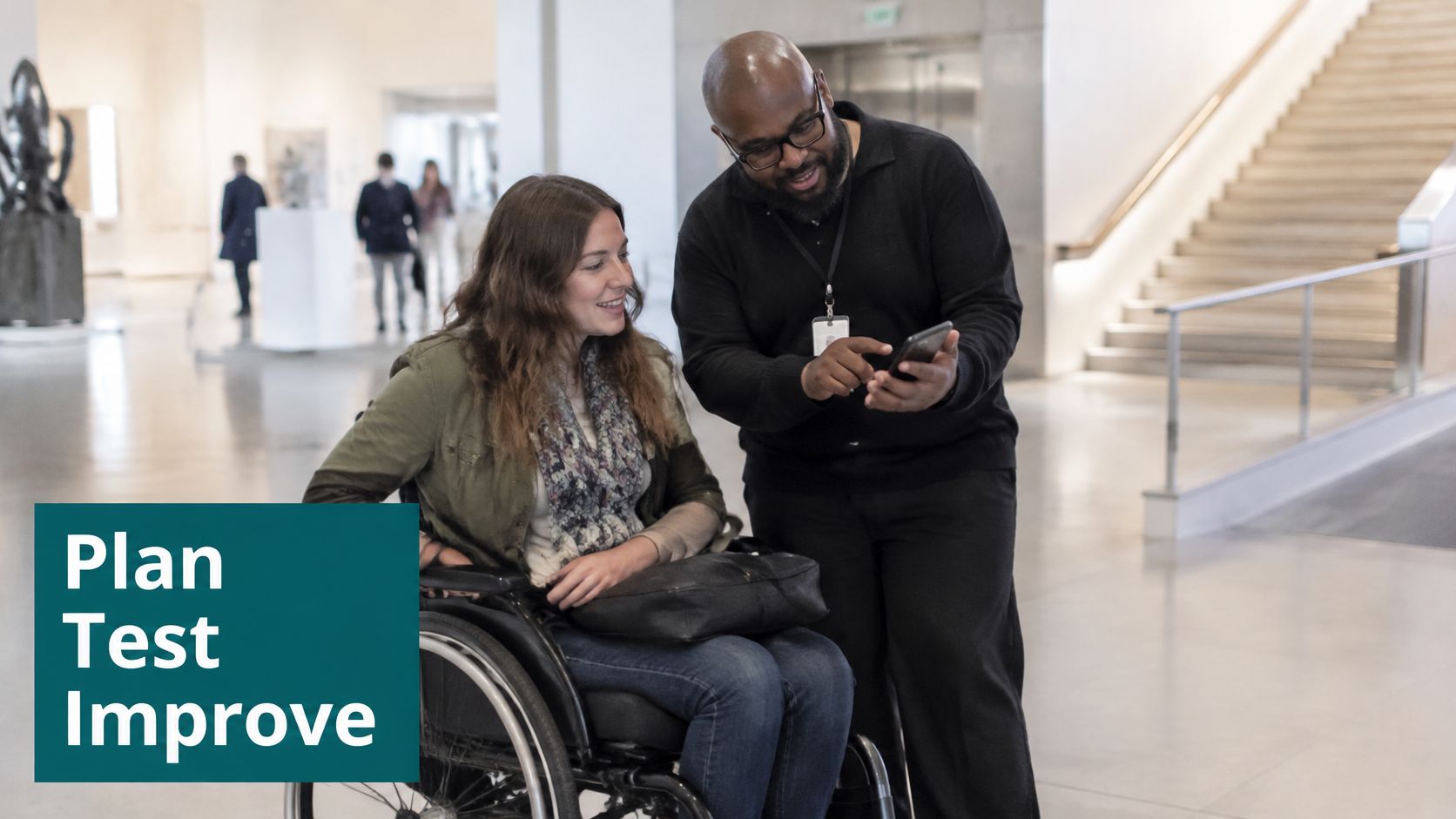 A man showing a smartphone to a woman in a wheelchair inside a large modern museum.