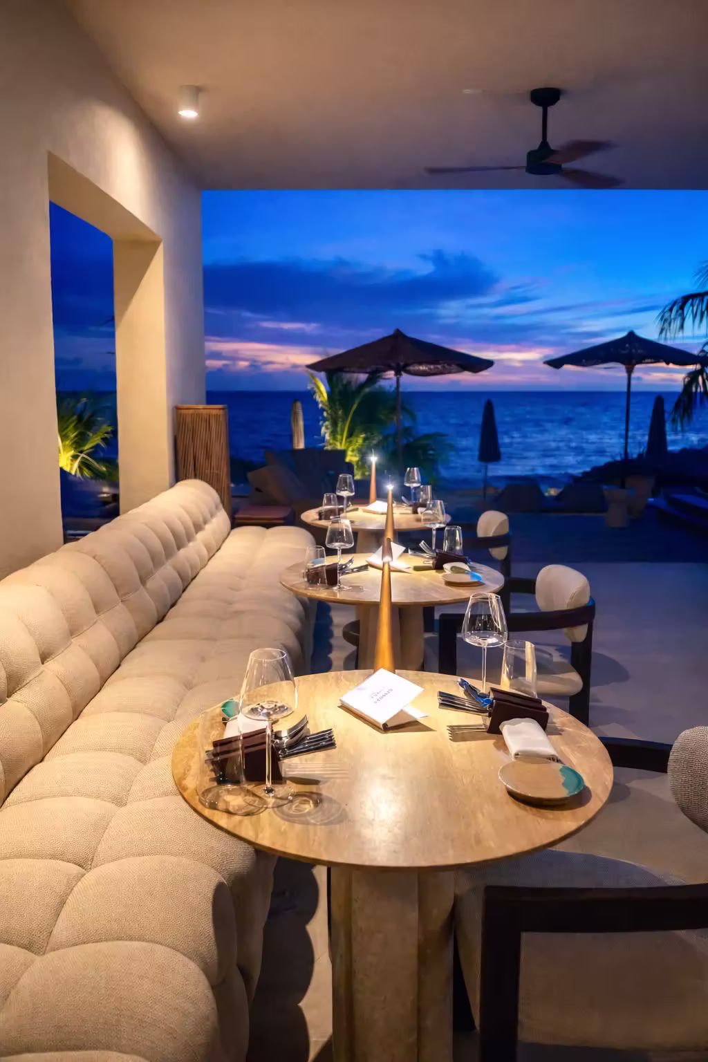 Elegant outdoor dining area at dusk with wooden tables, cushioned chairs, and ocean view under colorful sky.