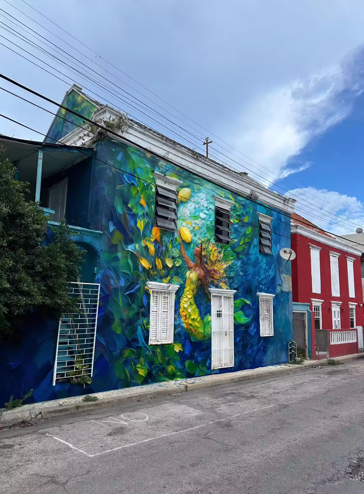 Street view of a building with a large vibrant mural of a yellow-green mermaid surrounded by underwater plants and a blue background.