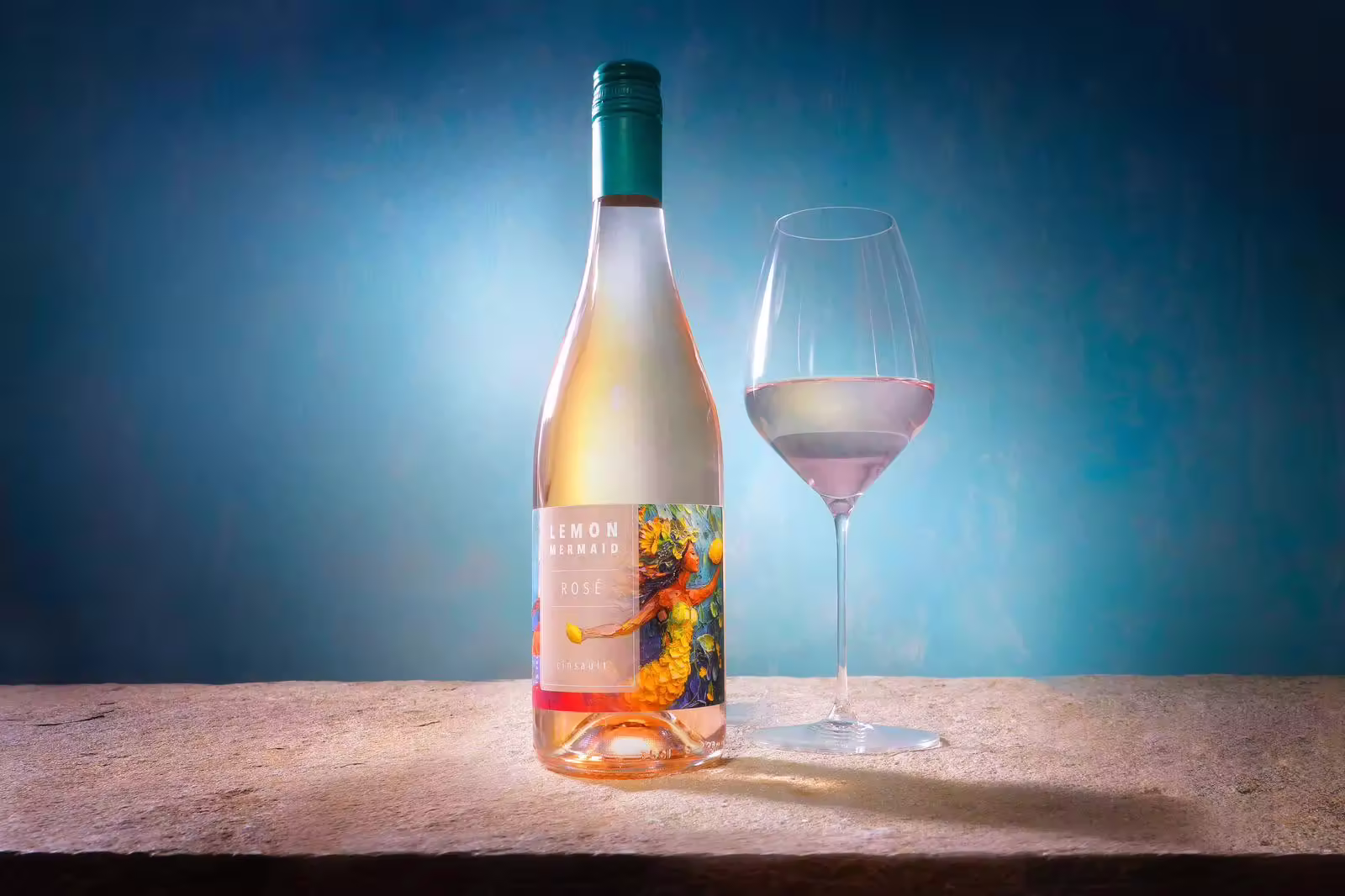 Bottle of Lemon Mermaid Rosé wine next to a wine glass filled with rosé on a stone surface with a blue gradient background.
