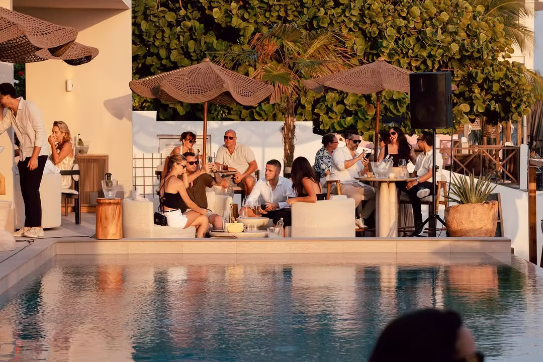 Group of people socializing outdoors near a pool with umbrellas and lounge seating during sunset.