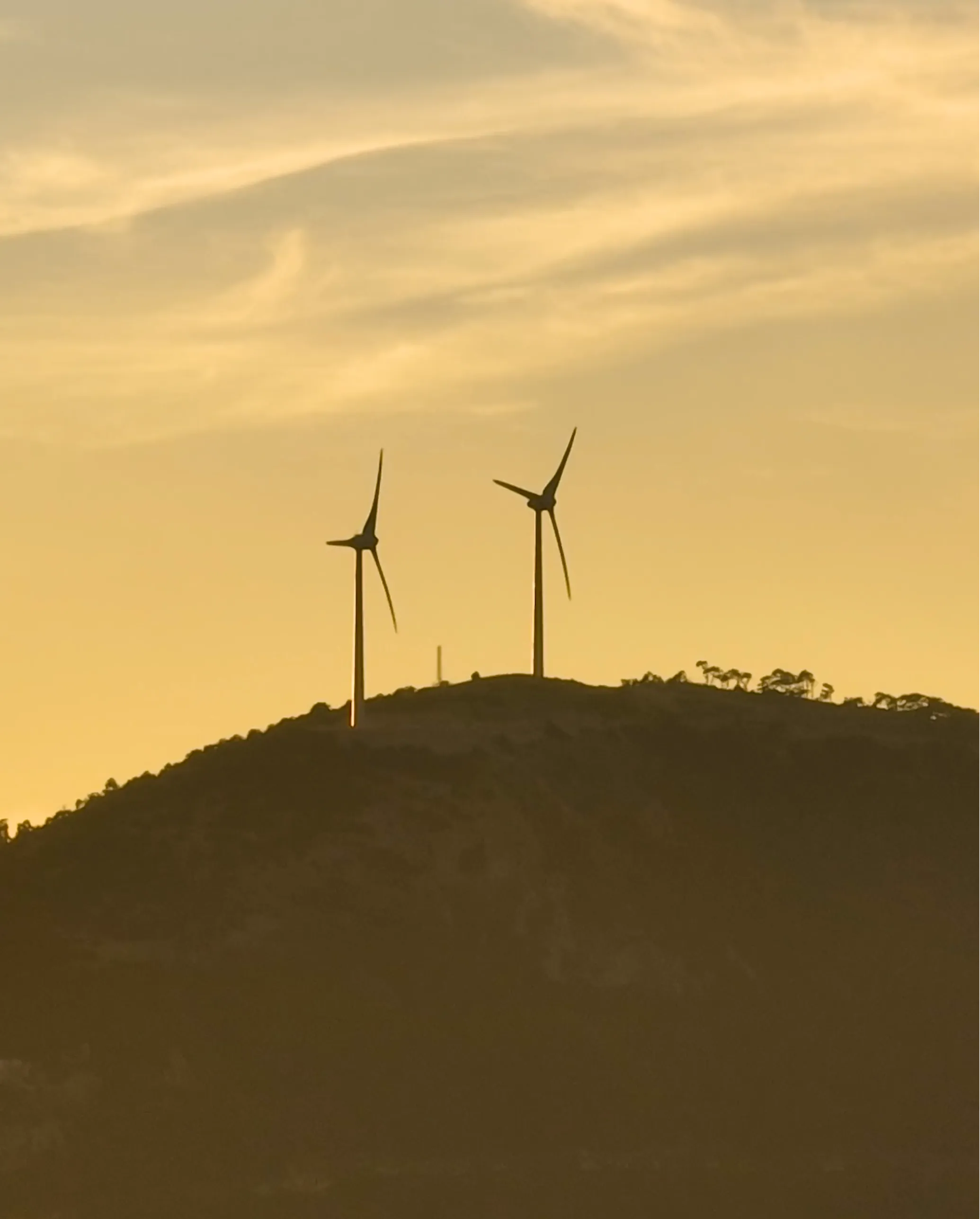 Two wind turbines on a hill silhouetted against a yellow-orange sunset sky with clouds.