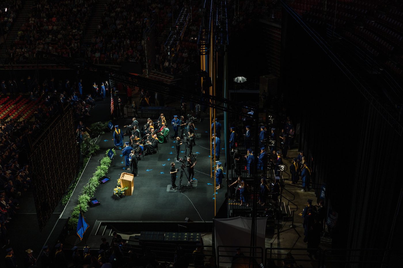 Arena commencement stage with graduates and camera crew at the podium