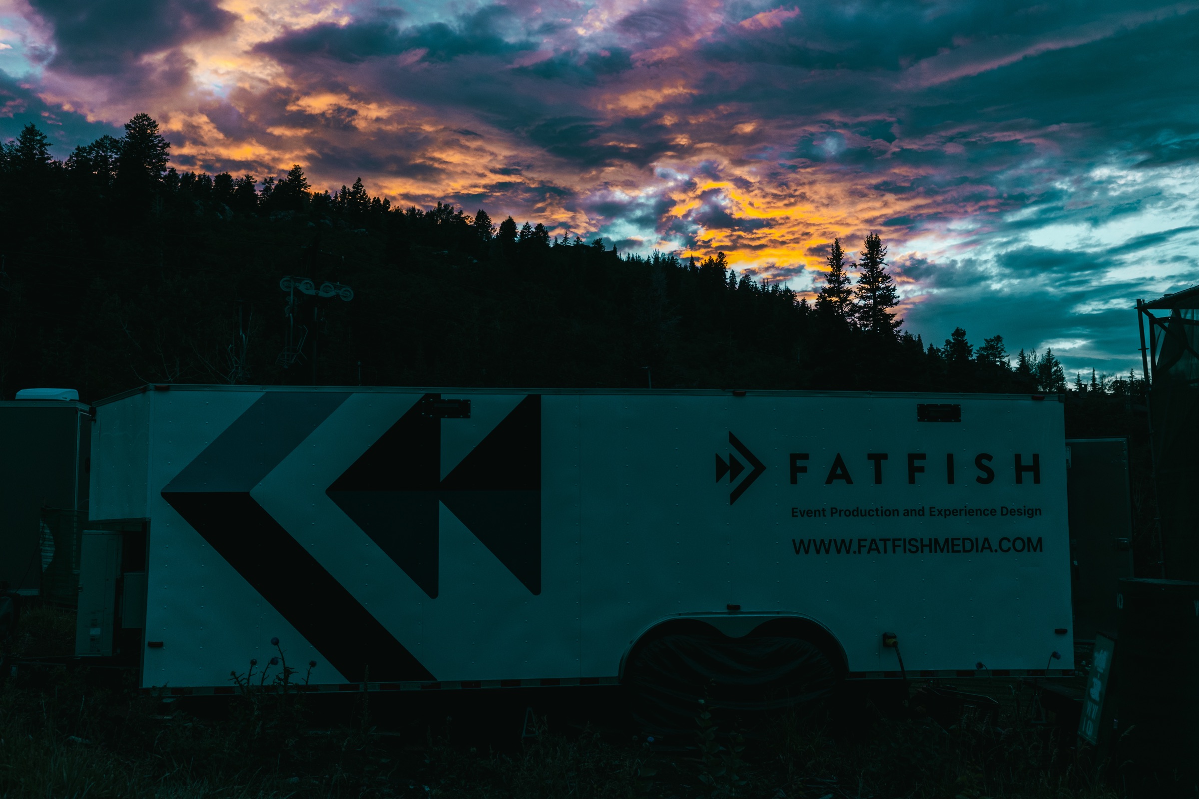 Small broadcast trailer silhouetted against a sunset over forested hills