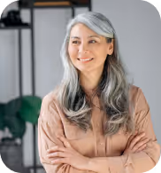 Mujer sonriente de cabello largo y canoso con los brazos cruzados, vestida con una blusa marrón claro, en un entorno interior moderno.