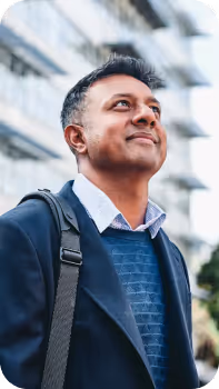 Hombre sonriente con abrigo azul y mochila mirando hacia arriba frente a un edificio moderno.
