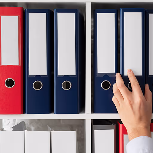 Mano tomando una carpeta azul de una estantería con varias carpetas azules y rojas organizadas.