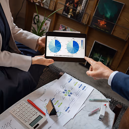 Dos personas revisan gráficos circulares en una tableta durante una reunión, con documentos, calculadora y bolígrafo sobre una mesa.