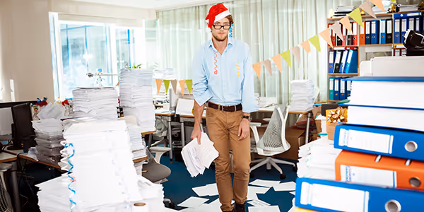 Hombre con gorro de Santa en oficina llena de montones de papeles y archivadores apilados por todas partes.