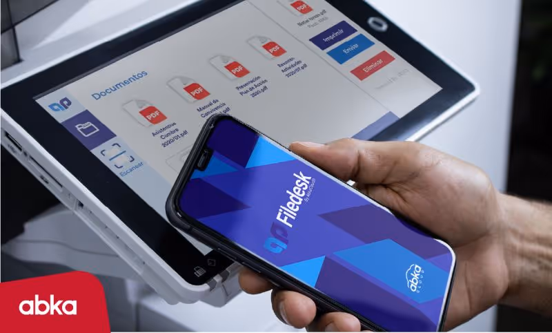 Mano sosteniendo un teléfono móvil con la pantalla que muestra la aplicación Filedesk frente a una pantalla táctil con documentos PDF listados.