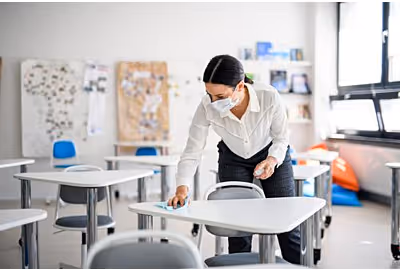 Person wearing a mask cleaning a desk in a classroom with chairs and educational posters.