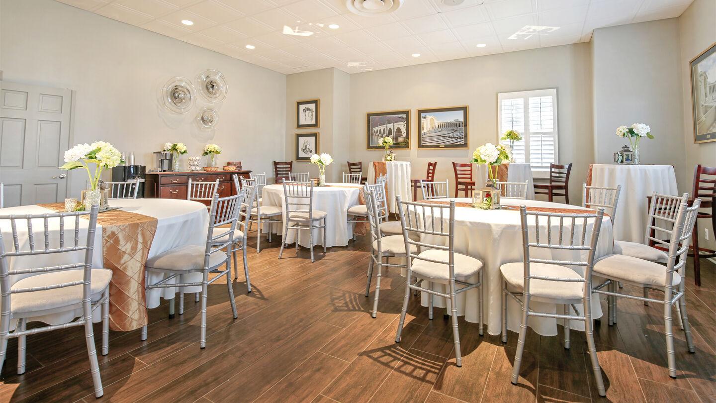 Bright, elegant event room with round tables covered in white and peach tablecloths, silver chairs, and floral centerpieces.