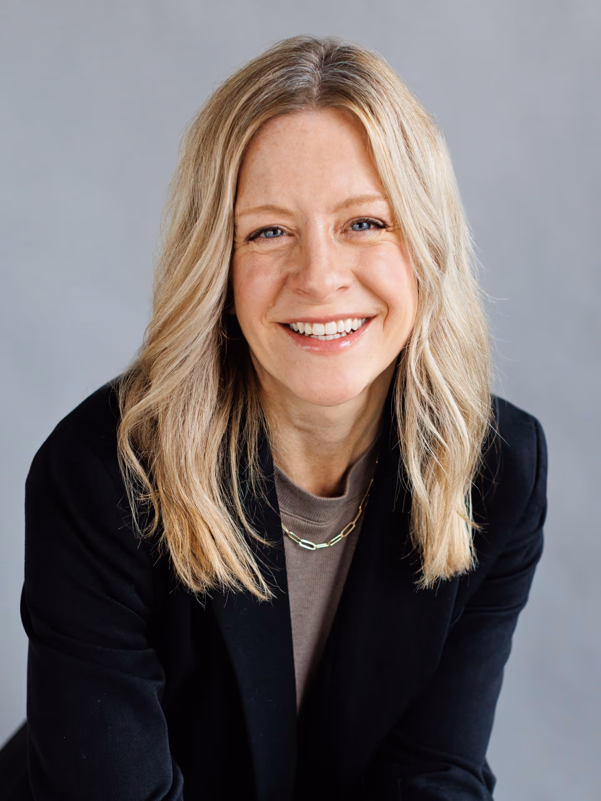 Smiling blonde woman with wavy hair wearing a black blazer and gold chain necklace against a gray background.