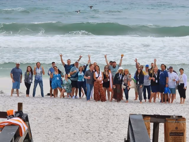 Group of people standing on a sandy beach near the ocean, smiling and waving at the camera under a cloudy sky.