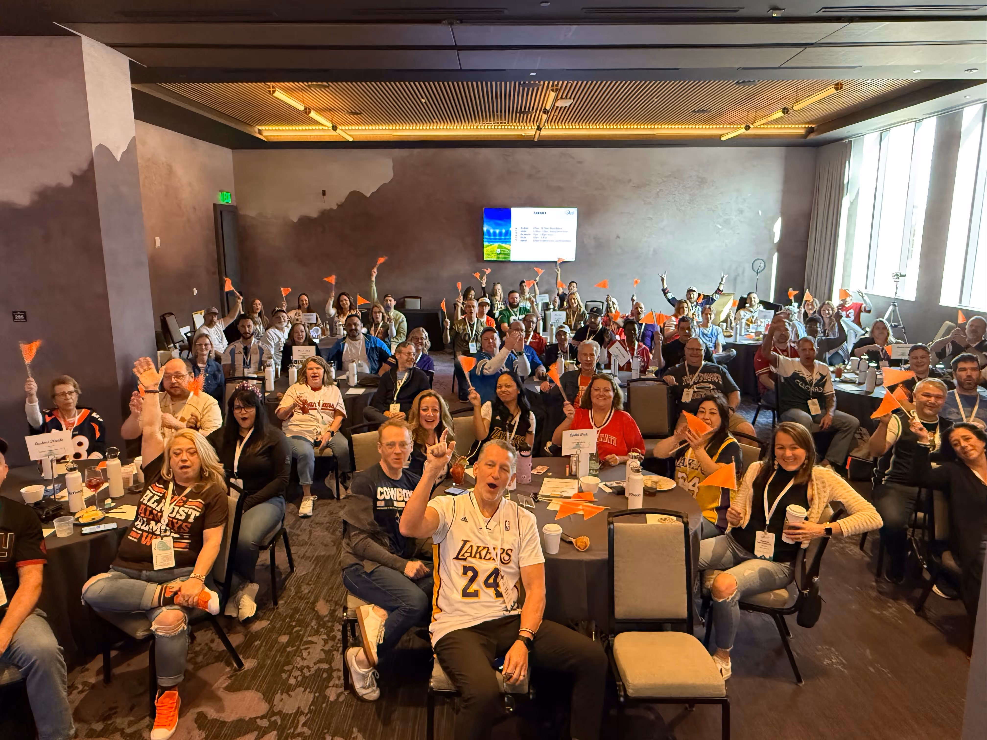 Large group of people sitting at tables in a conference room waving small orange flags and smiling.