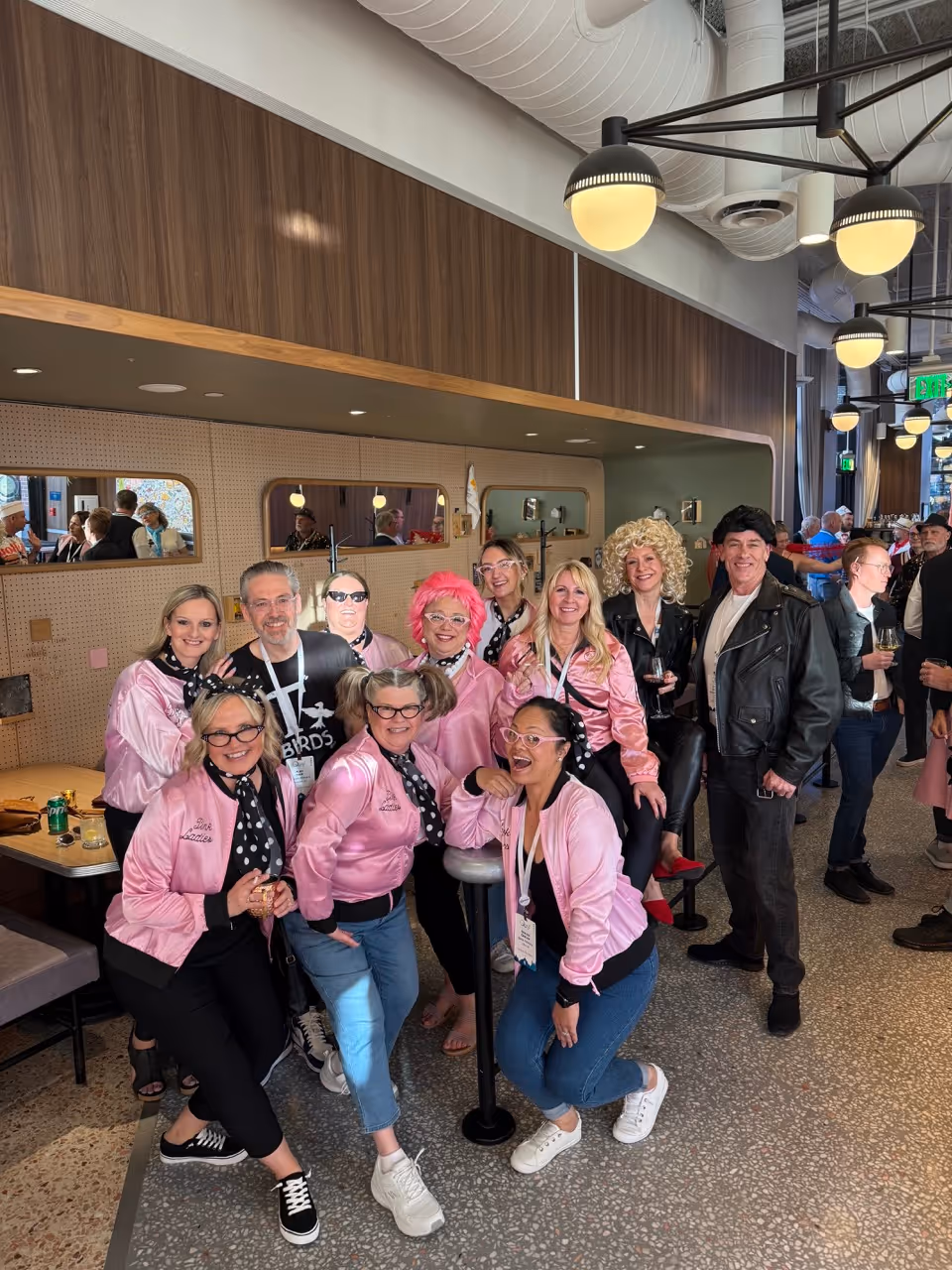 Group of people in retro pink jackets and 1950s style outfits posing indoors at a social event.