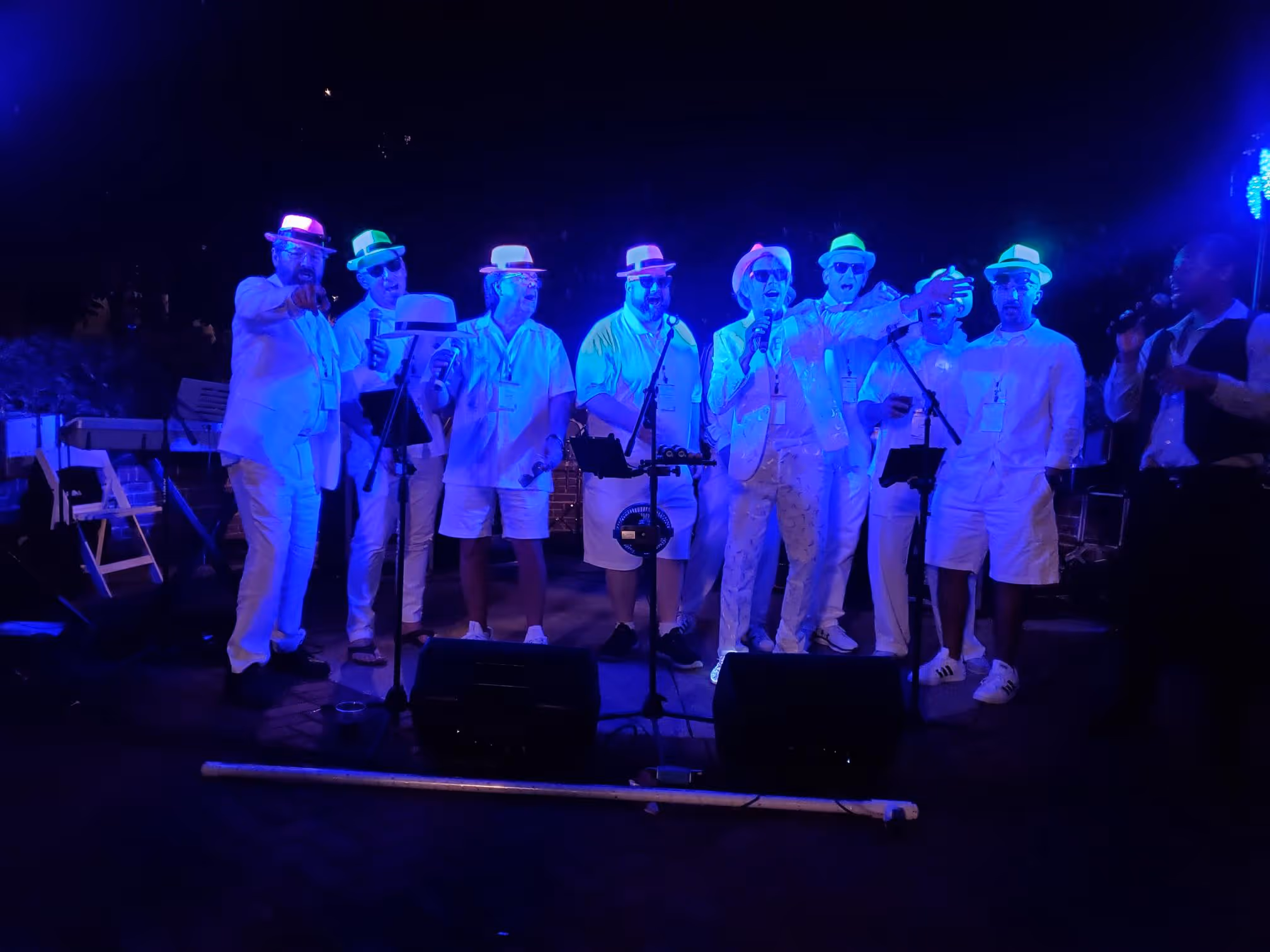 Group of men wearing white clothing and colorful hats singing on stage under blue lighting.