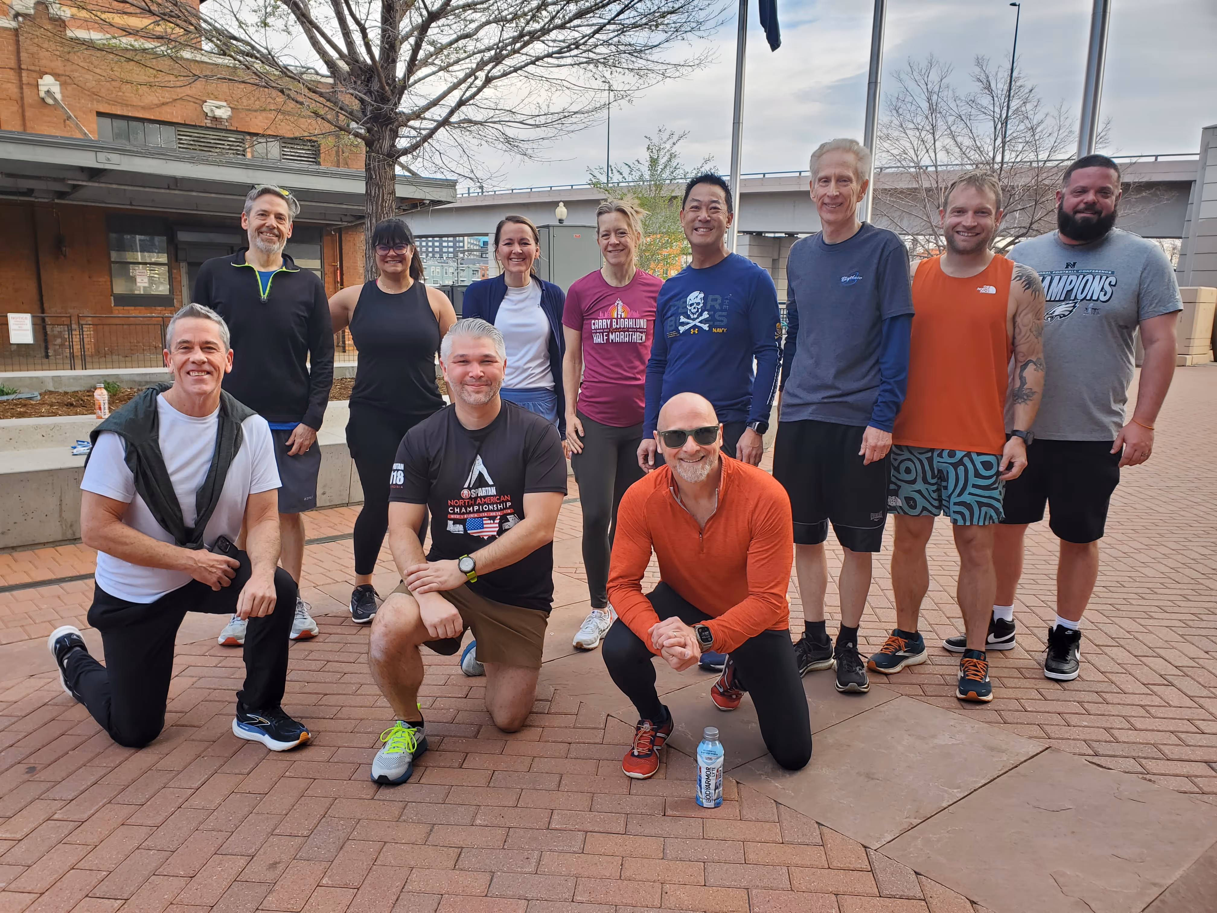 Group of twelve people outdoors posing on a brick pavement with some kneeling and others standing, with leafless trees and urban buildings in the background.