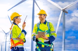 Two engineers in yellow safety helmets and high-visibility jackets discussing work near wind turbines.
