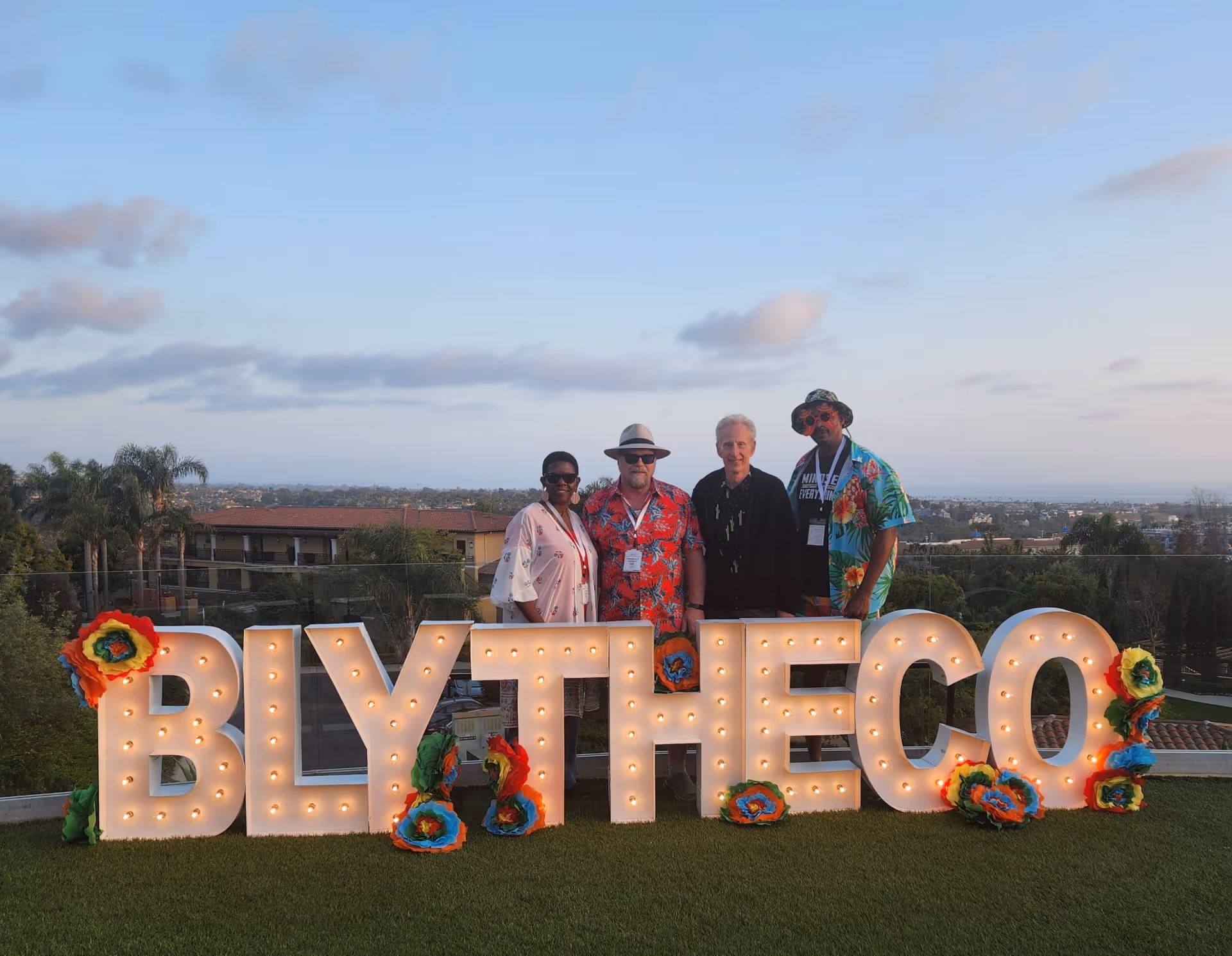 Four people standing behind illuminated letters spelling BLYTHECO decorated with colorful flowers on a grassy hill at sunset.