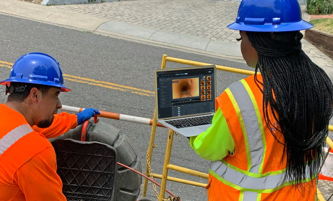 Two construction workers wearing blue helmets and orange safety vests monitoring a laptop screen beside a roadwork site.