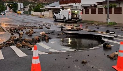 Large sinkhole on a street surrounded by debris and traffic cones with a white utility truck nearby.