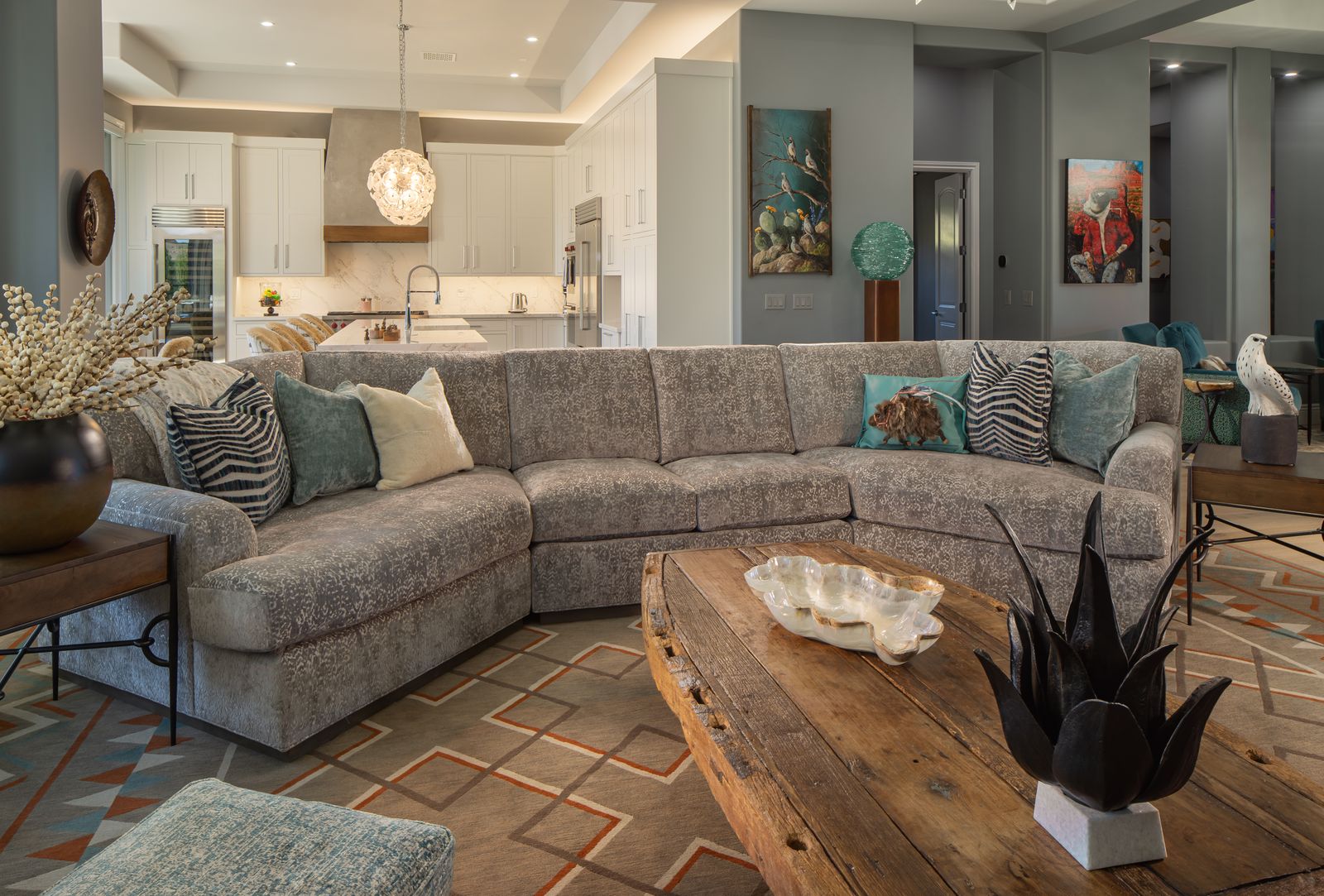 A modern living room with a large custom gray sectional sofa, wooden coffee table with decor, patterned rug, and an open kitchen in the background.