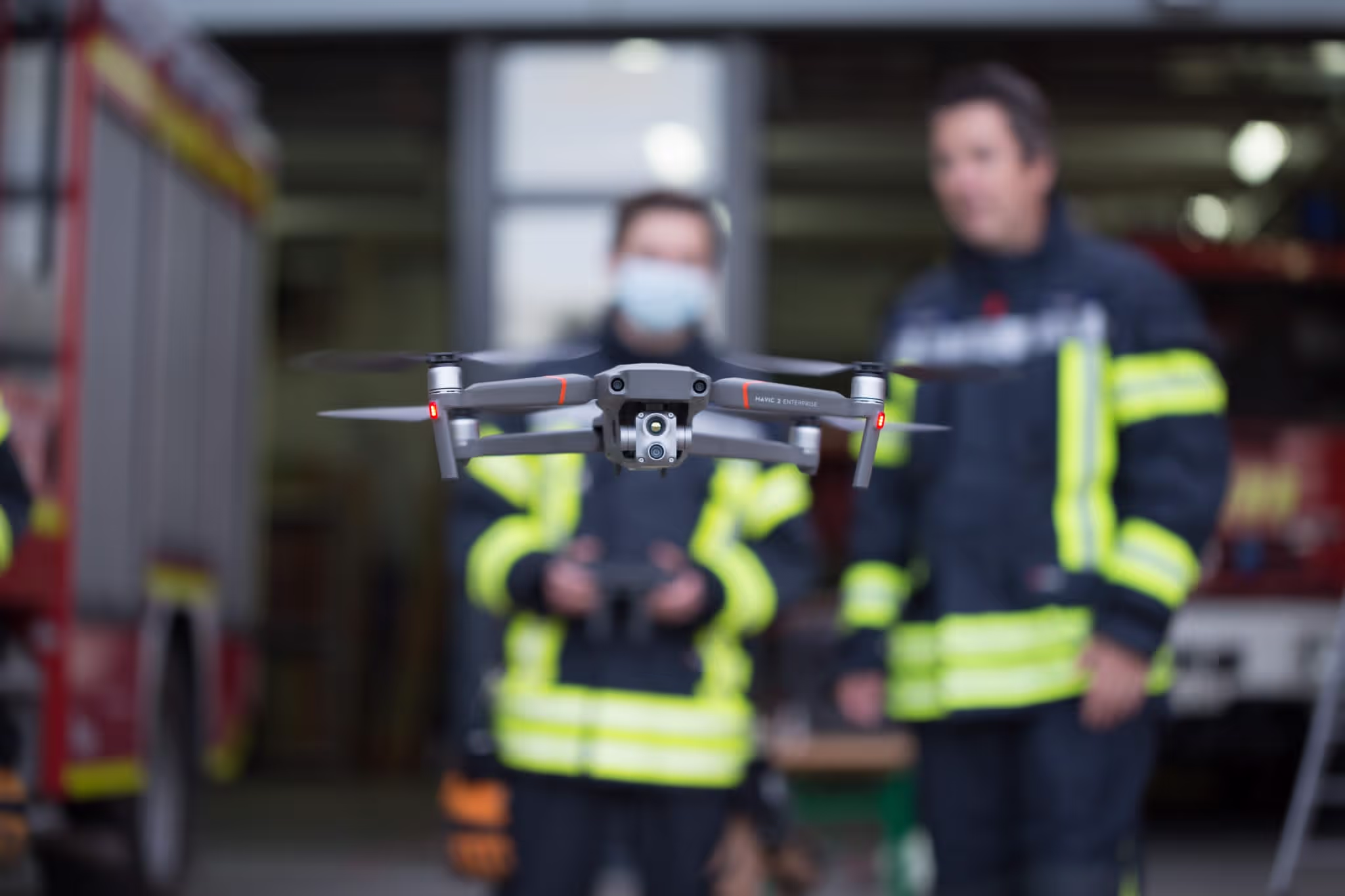 Eine Drohne schwebt im Vordergrund, während im Hintergrund zwei Feuerwehrleute in Einsatzkleidung stehen.